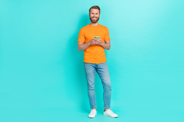 Fototapeta na wymiar Full size photo of boyfriend guy hold telephone wear orange t-shirt jeans sneakers isolated on teal color backgroiund