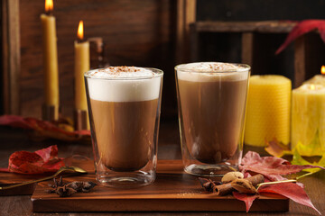 Two transparent glasses with cappuccino with black coffee, milk foam and cinnamon in autumn setting, burning candles, pumpkins, yellow and red tree leaves on wooden background