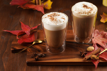 Two transparent glasses with cappuccino with black coffee, milk foam and cinnamon in autumn setting, burning candles, pumpkins, yellow and red tree leaves on wooden background