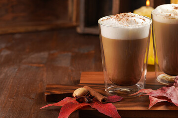 Two transparent glasses with cappuccino with black coffee, milk foam and cinnamon in autumn setting, burning candles, pumpkins, yellow and red tree leaves on wooden background