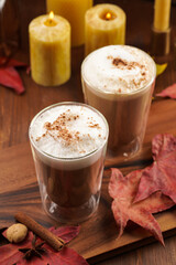 Two transparent glasses with cappuccino with black coffee, milk foam and cinnamon in autumn setting, burning candles, pumpkins, yellow and red tree leaves on wooden background