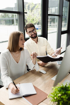 Smiling businessman holding paper folder near intern writing on notebook and computer in office.