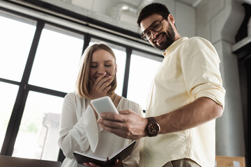 Low angle view of cheerful intern with notebook looking at smartphone near businessman in office.