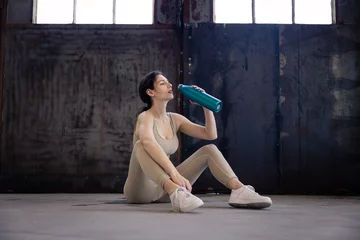 Tuinposter young woman in sportswear sits on the floor drinks from a bottle in her hand and relaxes after workout in the gym, trainer cooldown with water after exercise © alexfotobar