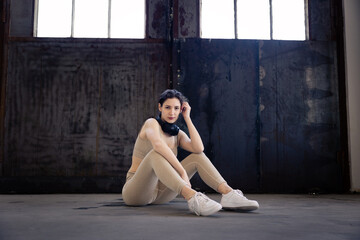 Fototapeta na wymiar young woman in sportswear sits on the floor with black headphones around her neck and looks at the camera on dark background