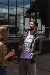 Cheerful businessman holding laptop and lunch box near colleague with coffee to go outdoors.