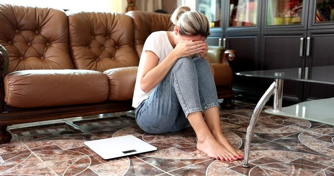 Woman is crying sitting on floor next to scales for measuring the maasa of body