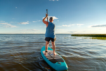 A man in shorts standing on a SUP board with a paddle floats on the water in the rays of the setting sun.