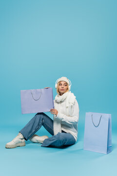 full length of blonde woman in winter outfit and wireless headphones sitting and holding shopping bag on blue.