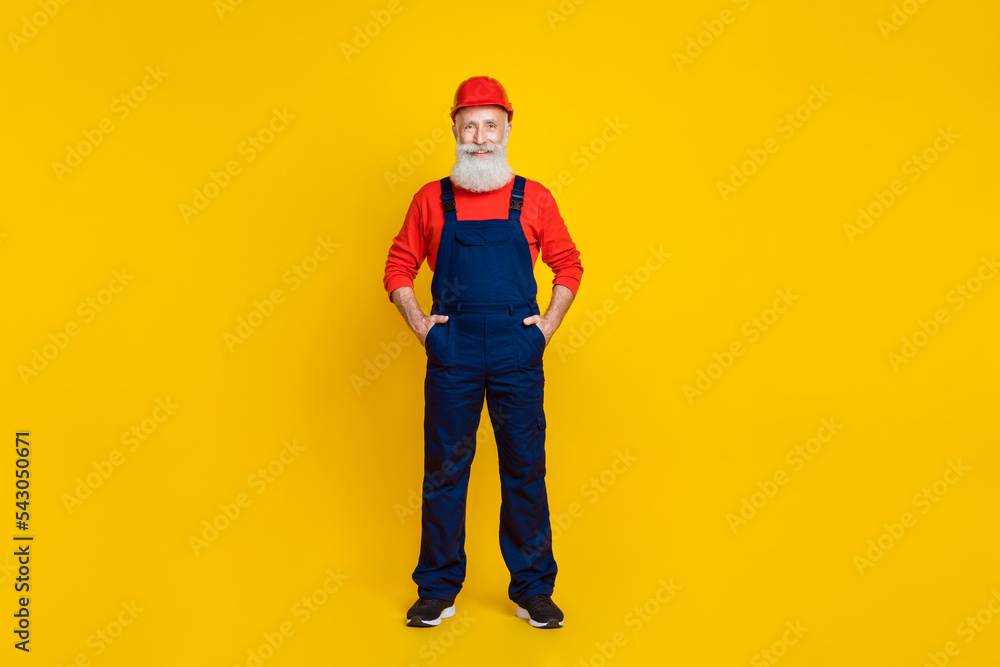 Wall mural full length photo of good mood senior man builder with white beard red helmet hands in pockets isola