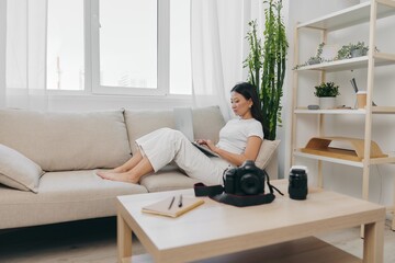 Asian woman photographer lying at home on the couch and reviewing photos from the camera in laptop for color correction retouching, work as a freelance business photographer for yourself home office