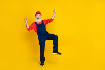 Fototapeta na wymiar Photo of positive funny senior guy dressed uniform overall red hardhat rising fists empty space isolated yellow color background