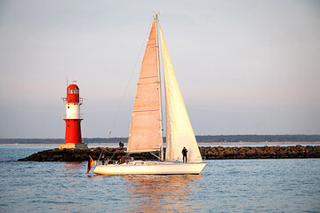 A sailing boat entering the harbor in front of a red lighthouse on the Baltic Sea coast.