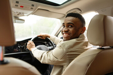 Back view shot of handsome african man sitting in car and holding steering wheel smiling happily at...
