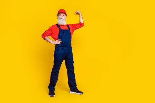 Photo of serious strong senior guy dressed uniform overall red hardhat rising fist empty space isolated yellow color background