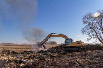 Close up view of a heavy equipment excavator moving trees and wooden debris into a fire pit near...