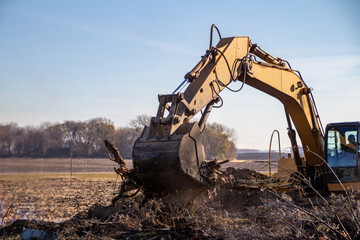 Close up view of a heavy equipment excavator moving trees and wooden debris into a fire pit near the area of a demolished old farm building