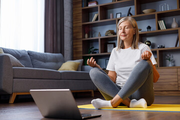 Gray-haired happy mature woman doing gymnastics and yoga at home online with laptop. Active and healthy lifestyle for pensioners. Fitness lessons online, recreation, well-being of the elderly.