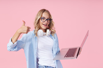 Portrait of a joyful teen girl in casual denim blue clothes with a laptop in her hands on a pink...