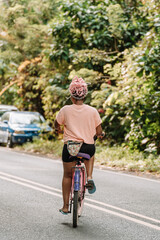 Imagen vertical desde atrás de una joven mujer afroamericana con cabello rosado andando en bicicleta por la calle en un día de verano. 