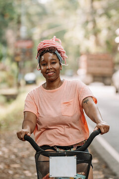 Imagen vertical de una hermosa y joven mujer afroamericana con cabello rosado andando en bicicleta al aire libre en un d&iacute;a de verano.  
