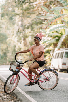 Imagen vertical de una joven mujer afroamericana con cabello rosado andando en bicicleta por la calle en un d&iacute;a de verano. 