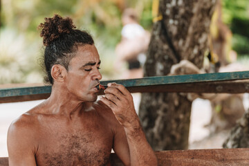 Imagen horizontal de un hombre adulto latino con cabello rizado sin camisa en la playa fumando hierba con una pipa en un día de verano.  - obrazy, fototapety, plakaty