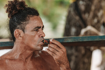 Imagen horizontal de un hombre adulto latino con cabello rizado sin camisa en la playa fumando hierba con una pipa en un día de verano.  - obrazy, fototapety, plakaty