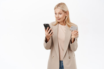 Online shopping. Smiling blond elegant woman looking at mobile phone, holding credit card, paying in application, standing over white background