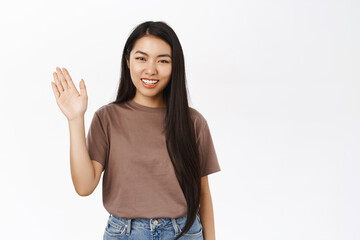 Friendly korean girl waves hand, say hello, hi and smiles at you, greets, stands over white studio...