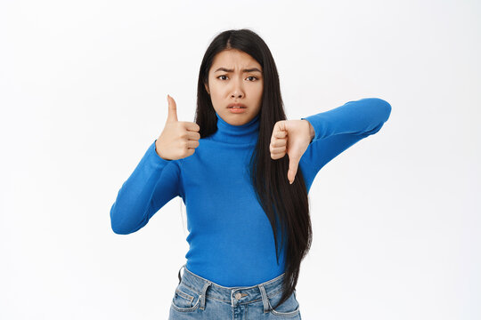 Like or dislike. Sad korean girl shows thumbs up and thumbs down, complicated with choice, dont know what to do, standing over white background