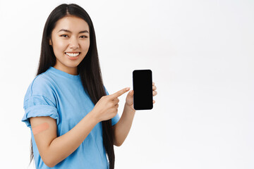 Smiling asian girl with band aid after coronavirus vaccine, shows smartphone screen, covid health certificate on mobile phone, white background