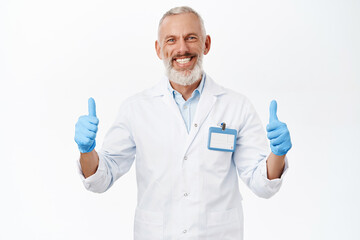 Smiling senior doctor, medical worker showing thumbs up, recommending clinic, standing over white background