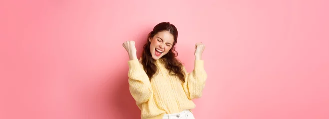 Fotobehang Girl screams with joy and fist pump, say yes, achieve goal or success, celebrating achievement, triumphing and winning, standing over pink background © Mix and Match Studio
