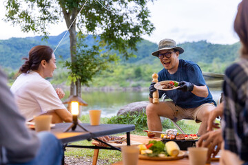 Group of Asian people friends enjoy outdoor lifestyle hiking and camping together on holiday travel vacation. Happy man cooking food grilled bbq and steak for dinner near the camp at summer sunset.