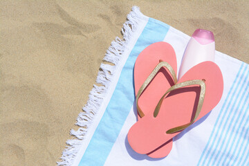 Striped towel with bottle of sunblock and flip flops on sandy beach, flat lay. Space for text