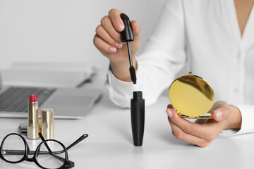 Young woman using cosmetic pocket mirror and mascara at white table indoors, closeup