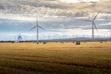 Windmills producing sustainable energy on the Alberta Prairies with round hay bales and a grain elevator with Canadian Rockies at background.