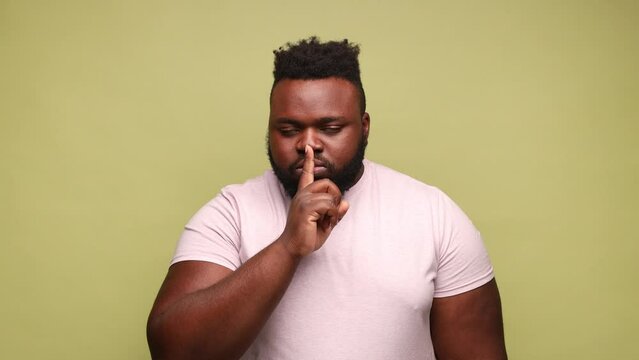 Serious African-american man wearing pink t-shirt holding finger on lips making hush silence gesture, asking to keep secret, don't speak. Indoor studio shot isolated on light green background.
