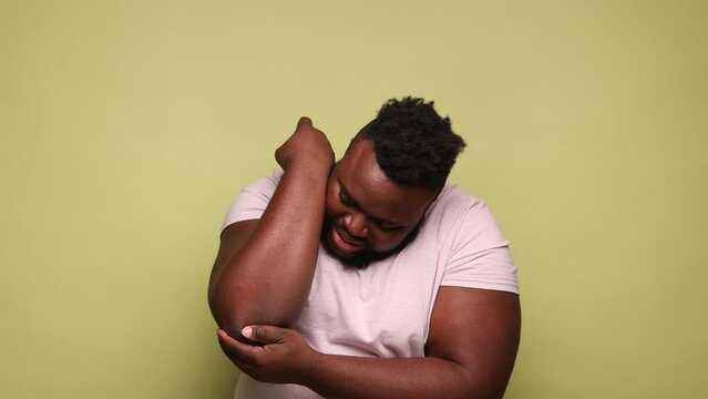 African-american man wearing pink t-shirt massaging aching elbow, frowning from acute pain, strain and strain problem, trapped nerves. Indoor studio shot isolated on light green background.