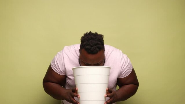 Young adult African-american sick bearded man wearing pink t-shirt having sudden vomit, suffering nausea, holding bin in hands. Indoor studio shot isolated on light green background.
