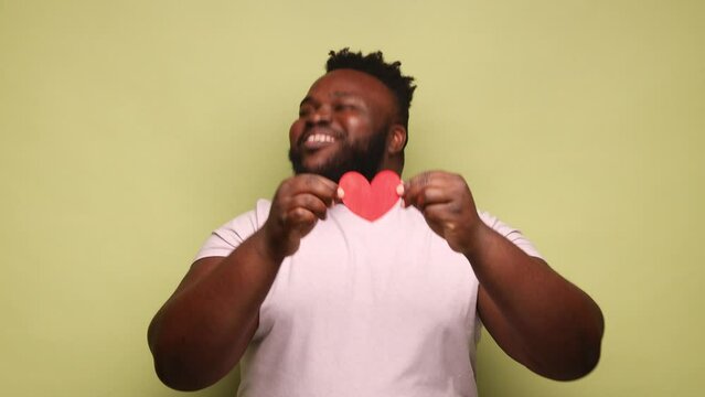 Extremely happy bearded African-american man wearing pink t-shirt dancing and showing little red heart, expressing love and happiness. Indoor studio shot isolated on light green background.