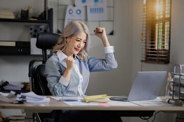happy asian businesswoman and showing winner gesture while sitting at workplace in office.