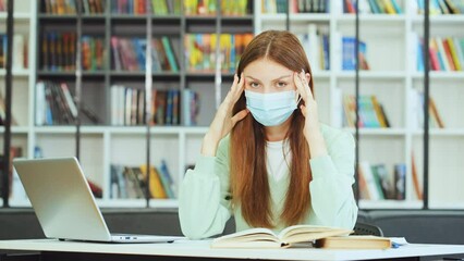 Young female student wearing protective mask working on laptop in library, suffering headache and starting massaging temples. Signs of illness
