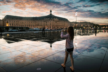 Young girl taking a photo of the famous Bordeaux water mirror with Place de la Bourse in the background at sunset - obrazy, fototapety, plakaty