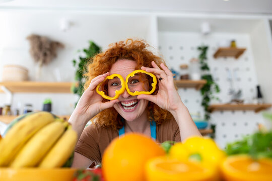 Photo of young woman smiling and holding pepper circles on her eyes while cooking salad with fresh vegetables in kitchen interior at home