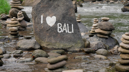 Stone towers on river rocks and large stone with inscription I love bali and painted heart in summer forest. Symbol of balance and harmony in tropical jungle. Zen symbol. Soft focus Film grain texture