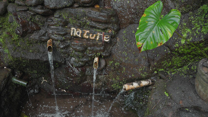 A tropical spring made by human hands using plastic pipes in rocks. a mountain spring with clean fresh water in with a tropical green leaf and inscription Nature. Film grain texture. Soft focus.
