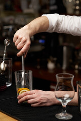 Cropped close-up shot of a bartender squeezing fresh juice out of an orange in a glass using a stainless steel muddler on the bar counter. Front view.