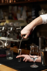 Cropped close-up shot of a bartender is pouring drink into a stainless steel jigger. Men's hands are pouring drink into a measuring cup on the bar counter. Front view.